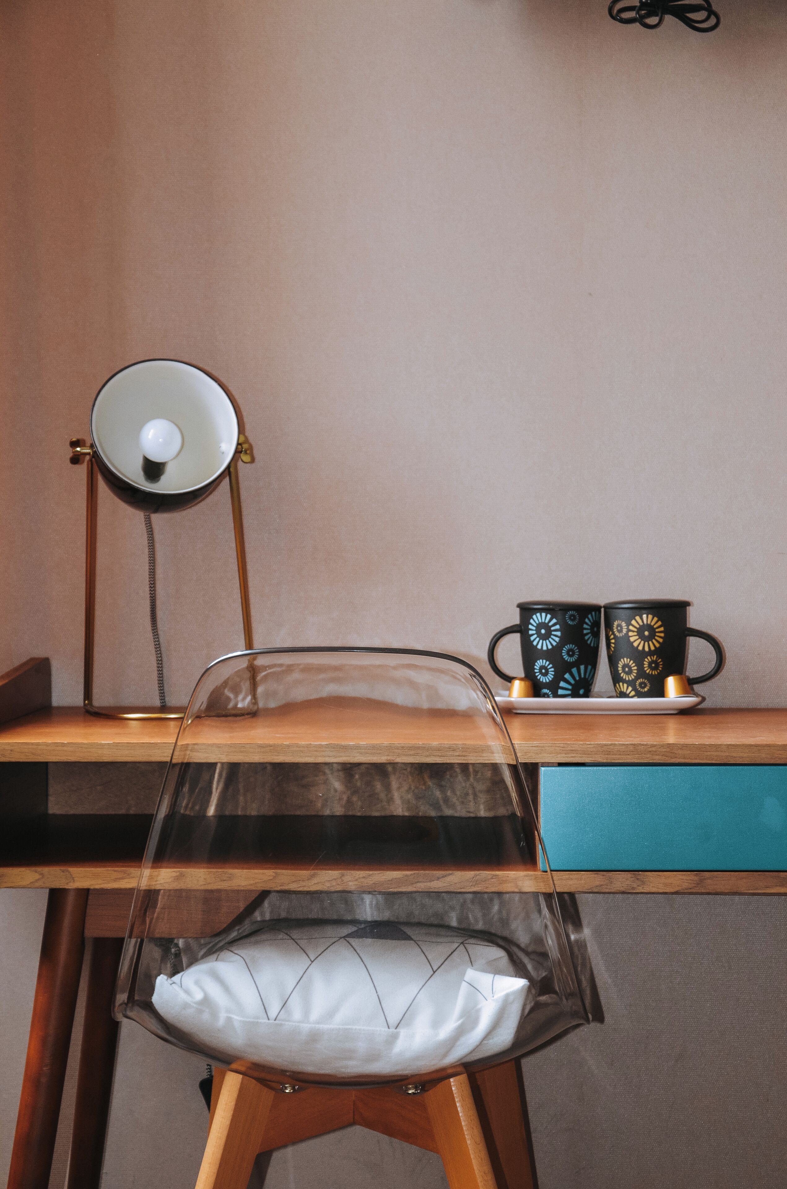 Une chaise transparente avec un coussin blanc, un bureau avec une lampe et deux tasses à motifs sur un plateau.
