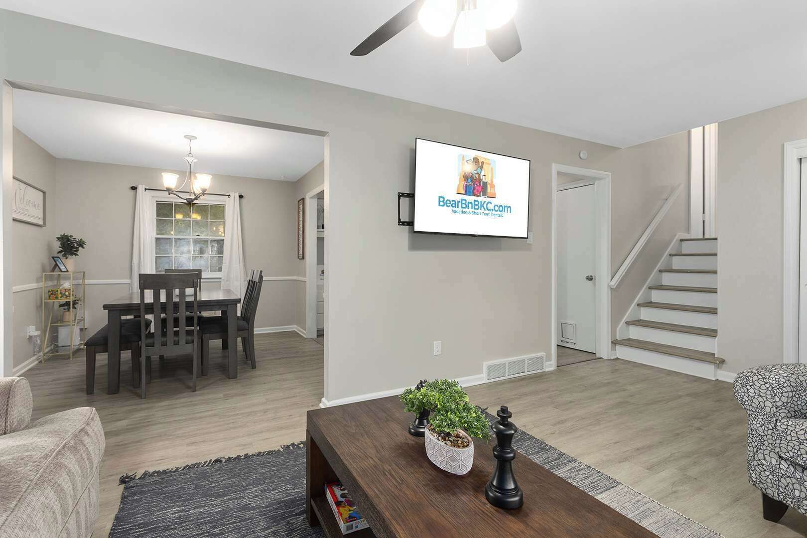 A modern living room with a dining area, a staircase, and a mounted TV displaying a website.