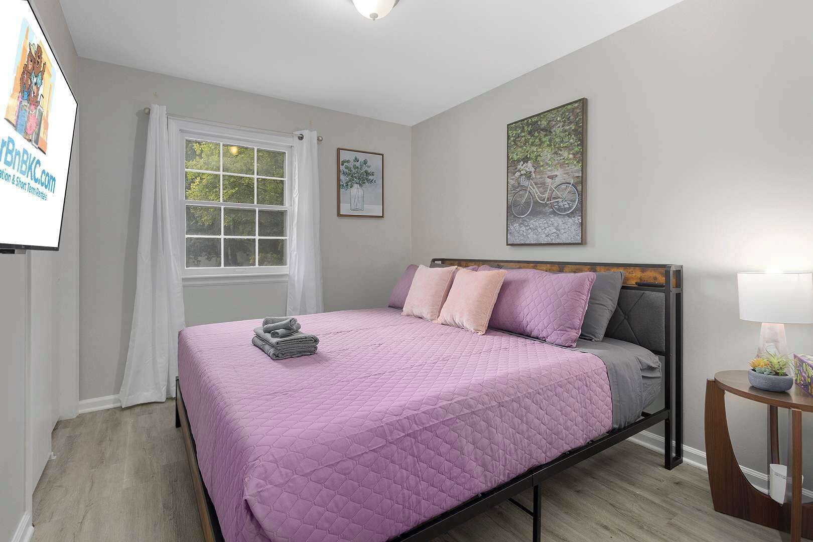 Un dormitorio con cama, ventana con cortinas, mesita de noche con lámpara y un cuadro en la pared.