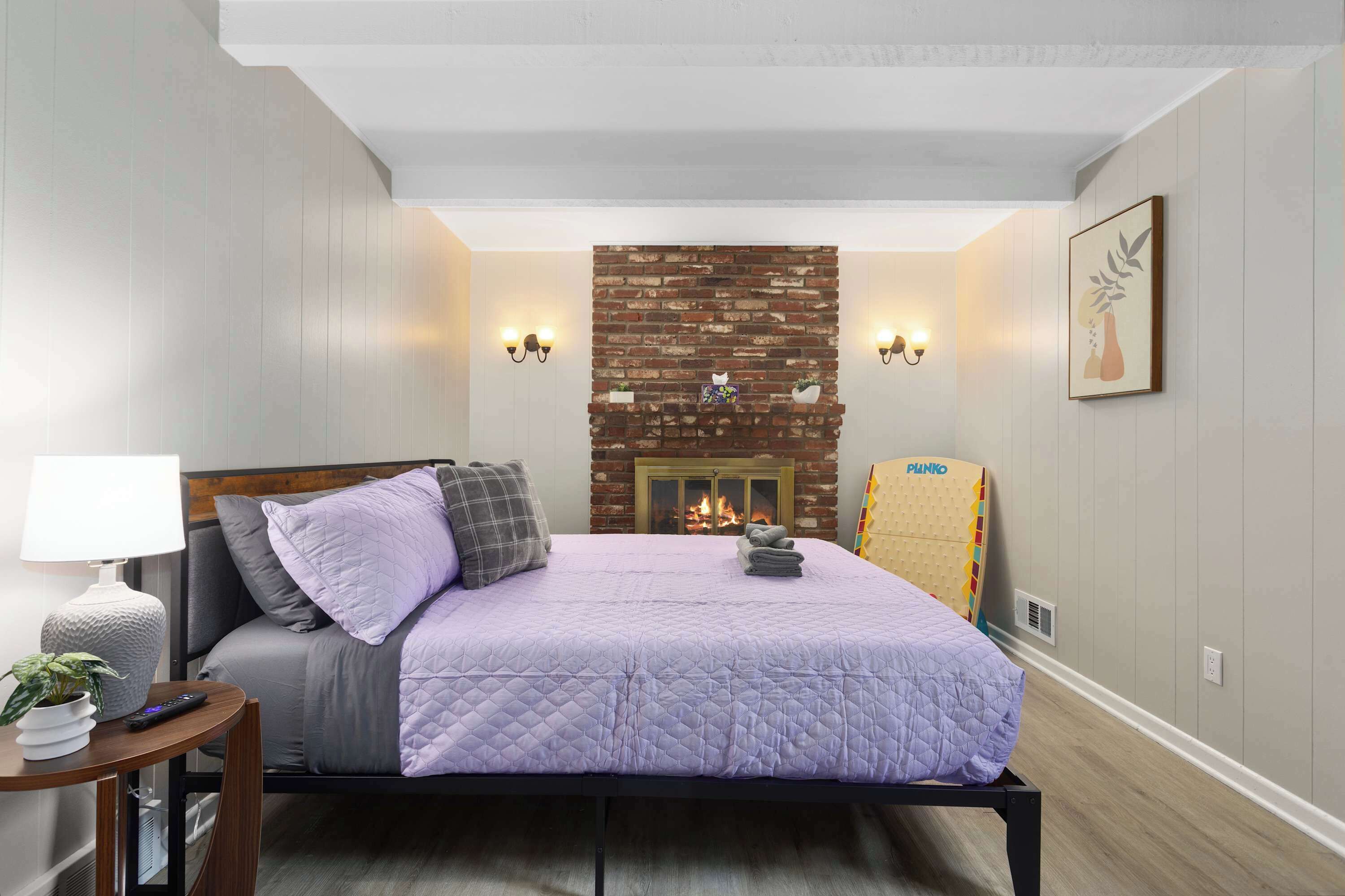 A bedroom with a bed, a nightstand, a lamp, a brick fireplace, and wall sconces.