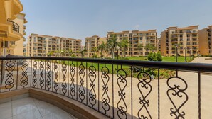 Un balcón con barandillas negras ornamentadas, suelo de baldosas y vista a edificios residenciales de varios pisos con balcones y vegetación.