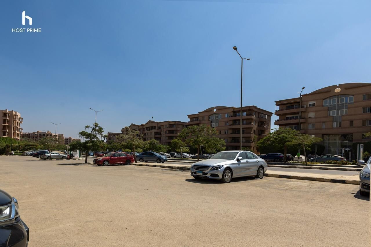 Un parcheggio con diverse automobili, tra cui una berlina bianca e un'auto rossa, di fronte a una fila di edifici a più piani con balconi.