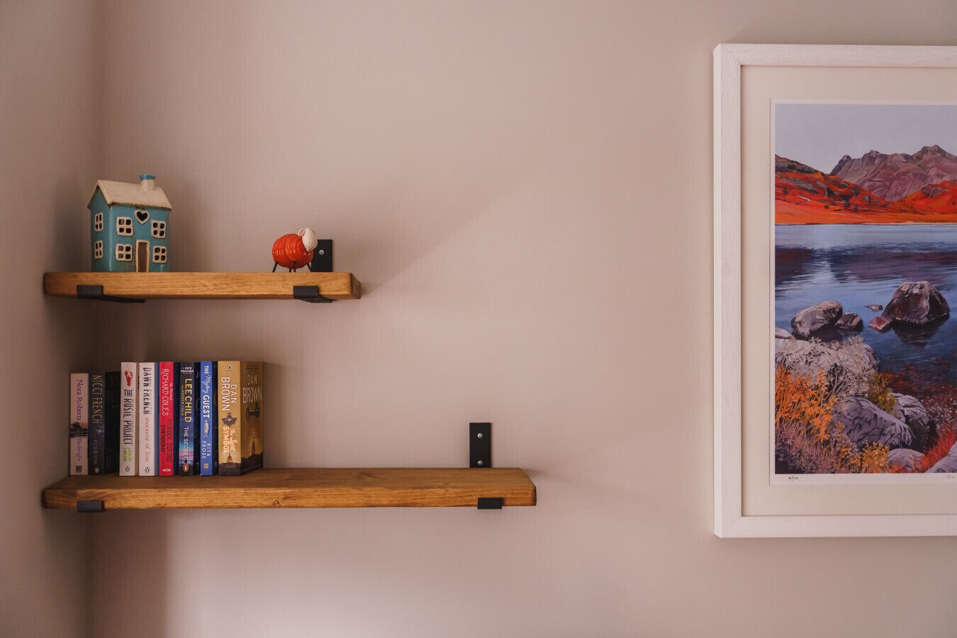 Twee houten planken met boeken en een miniatuurmodel van een huis, en een lijst met een landschapsdecoratie aan de muur.
