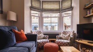 A cozy living room with a blue sofa, two patterned armchairs, a red ottoman, a wooden coffee table, a television, and a view of a blue building through large windows. - Herdwick Cottage, Stylish 3 bed, Central Ambleside (Ambleside)