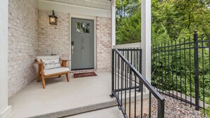 A porch with a brick wall, a wooden bench with a cushion, a door with a number, and a black metal railing.