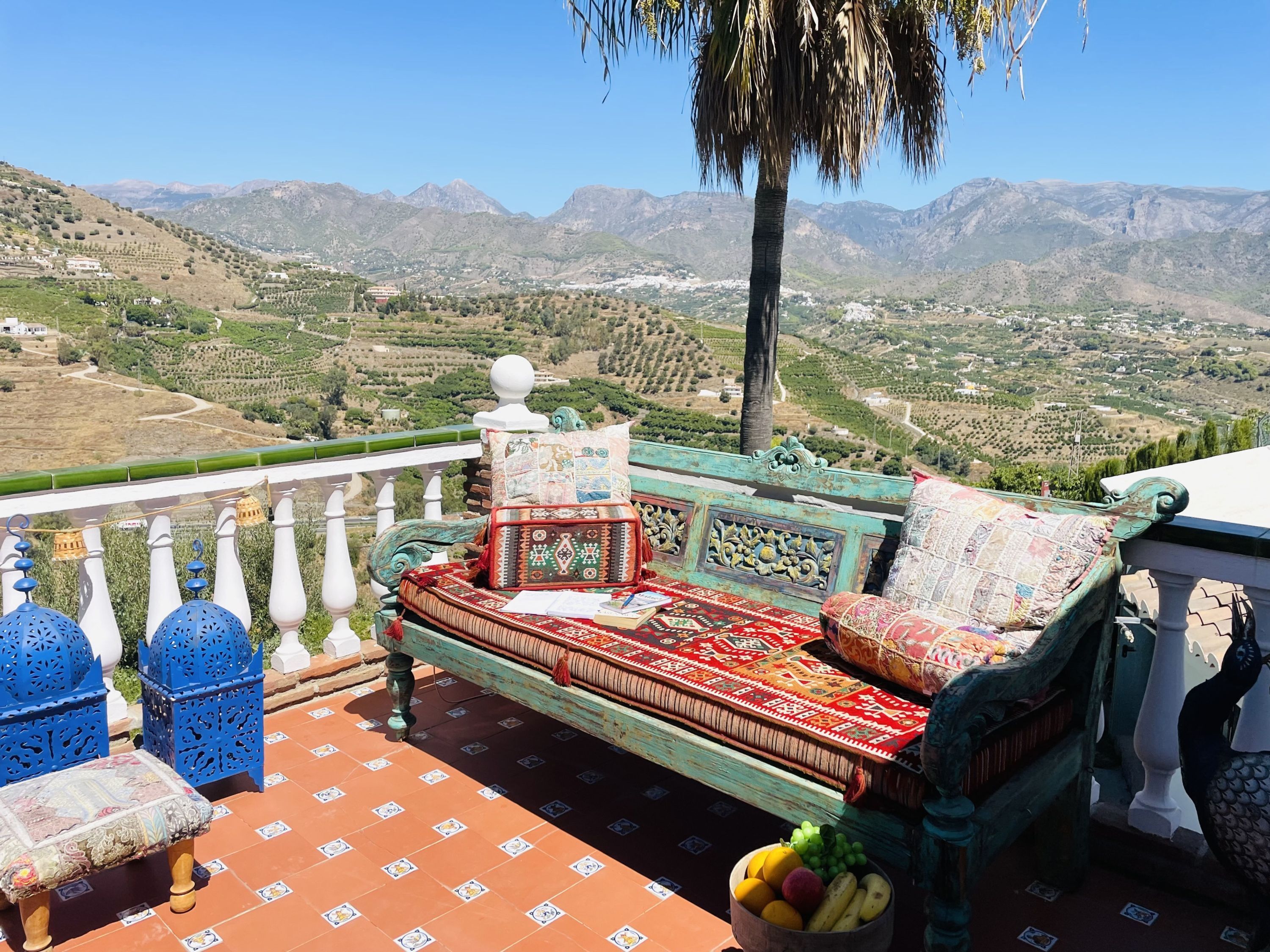 A balcony with a traditional sofa, a table with a bowl of fruit, and a view of a mountainous landscape.