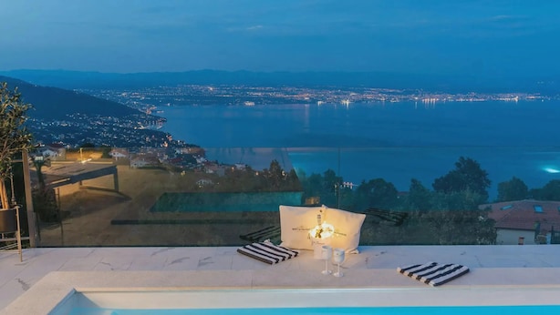 Vista da piscina com um horizonte urbano ao fundo, área de estar com um sofá e uma mesa, e céu claro.