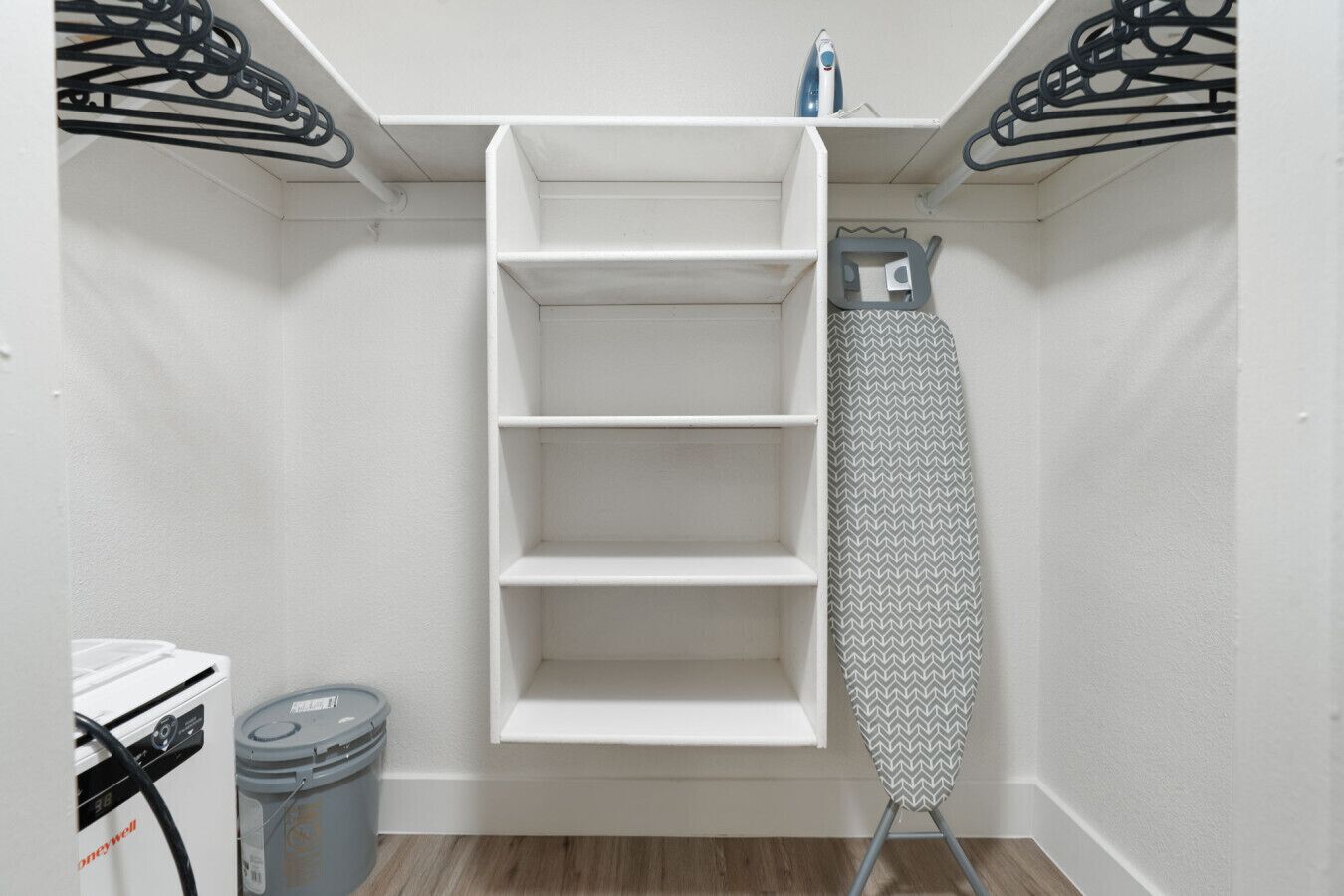 A laundry room with a white shelving unit, an ironing board, and a trash can.