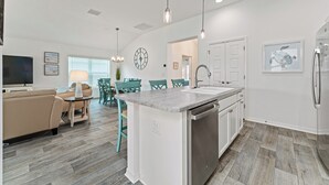 Une cuisine moderne dotée d’un îlot central, de meubles blancs et d’un lave-vaisselle encastré. On y trouve un coin repas avec des chaises et une table de couleur turquoise, ainsi qu’un salon avec un canapé beige et un téléviseur à écran plat.