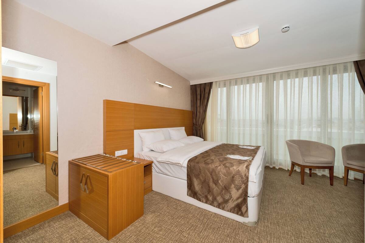 Habitació d'hotel amb un llit, una tauleta de nit de fusta, un mirall, una cadira i una finestra amb cortines.