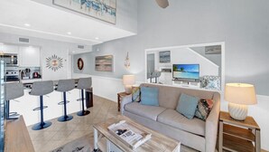 Un salon moderne avec un canapé, une table basse et des tabourets de bar. On y trouve un ventilateur de plafond, des œuvres d’art accrochées aux murs et, au fond, une cuisine.