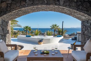 An outdoor seating area with a stone arch, a table with wine glasses and grapes, and a view of the sea.