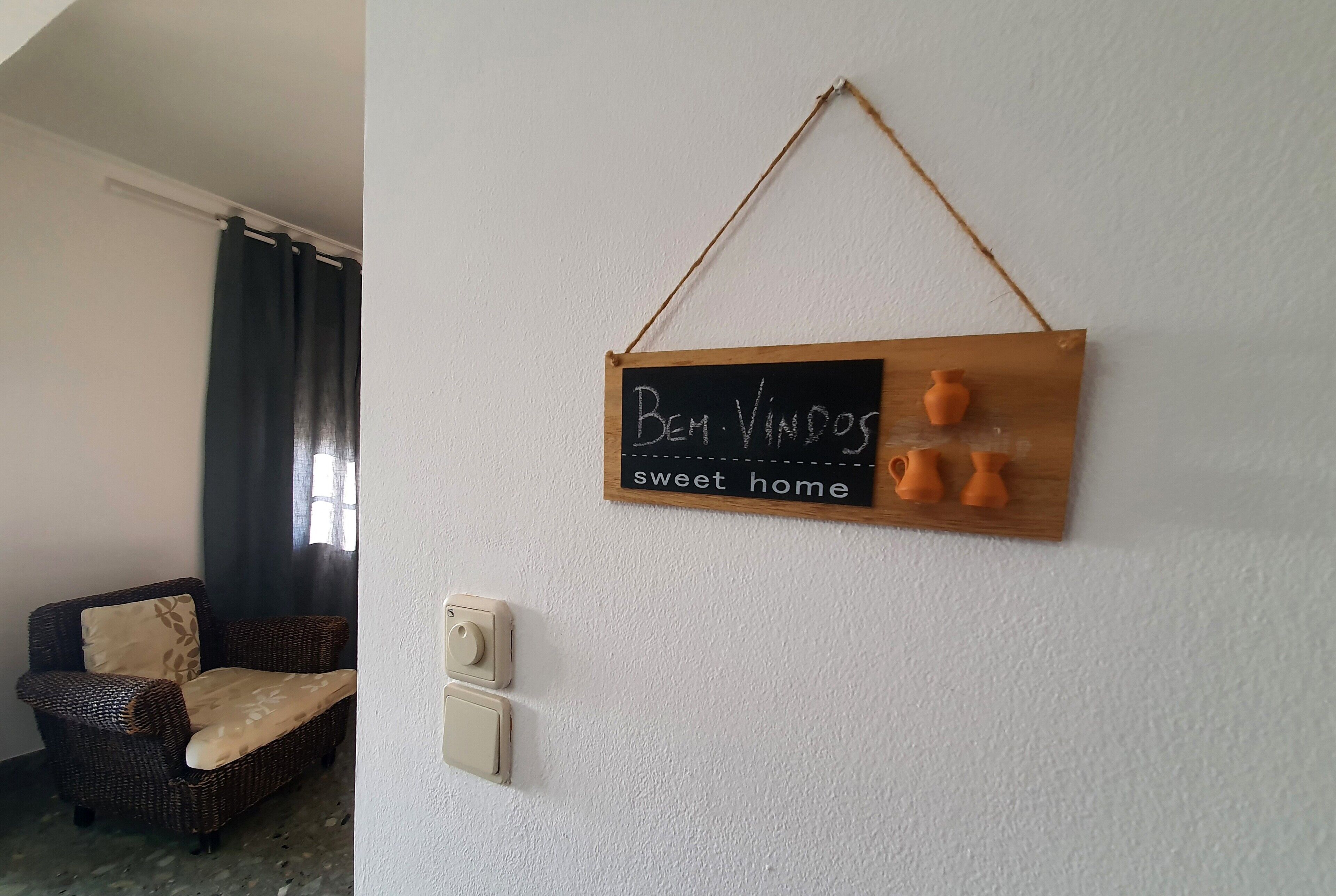 Et værelse med en fletstol, et ophængt skilt med teksten 'Bem Vindo' og 'hyggeligt hjem', samt en lyskontakt.