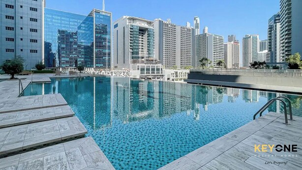A modern building complex with a pool area, featuring a clear blue pool, a tiled deck, and high-rise buildings in the background. - Majestine Stylish Studio with Canal View (Dubai)