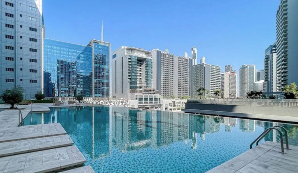A modern building complex with a pool area, featuring a clear blue pool, a tiled deck, and high-rise buildings in the background.