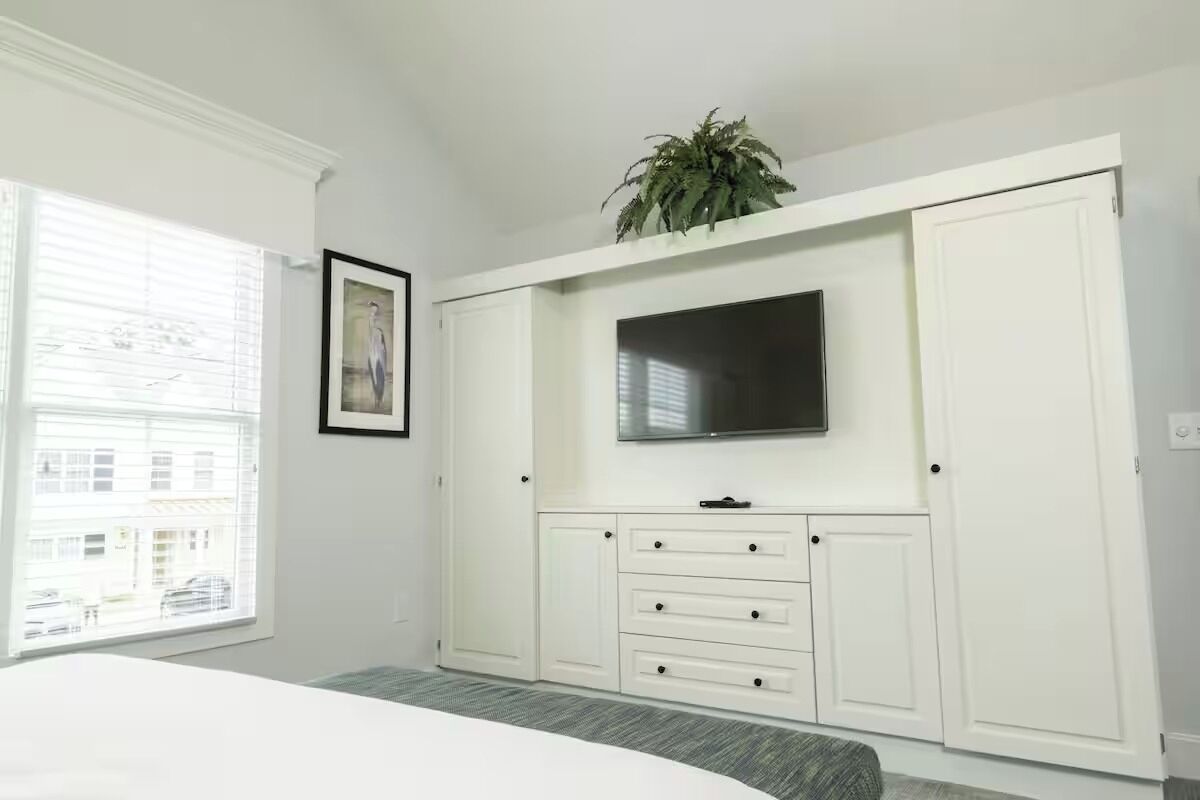 Une chambre à coucher avec un lit, un téléviseur fixé sur un meuble blanc, une fenêtre avec des stores et une plante posée sur le dessus du meuble.