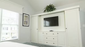 Un dormitorio con cama, televisor montado en un mueble blanco, ventana con persianas y una planta encima del mueble.