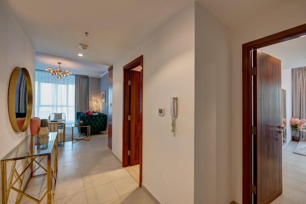 A modern hotel room with a large mirror, a wooden door, and a view into another room with a green sofa and a chandelier.