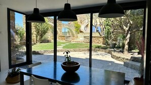 A dining area with a large table and chairs, a bowl of fruit, and a view of a stone building and garden through large windows. - Le Clos du Vialle (Grillon)