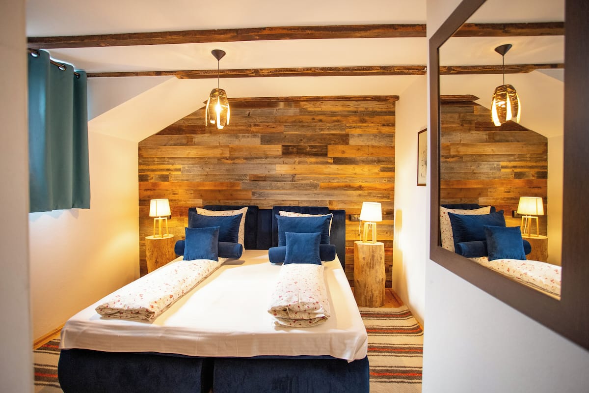A bedroom with a wooden headboard, a bed with blue bedding, two bedside lamps, and a mirror reflecting the room.
