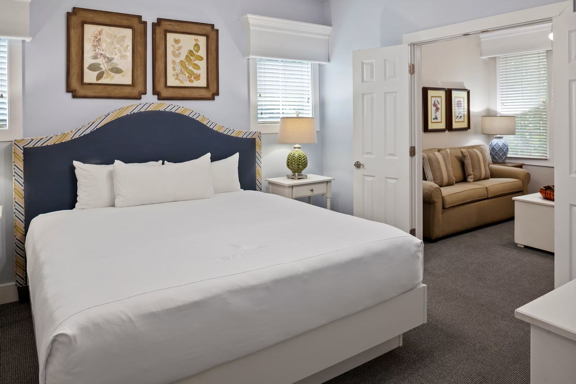 A hotel room with a large bed, a bedside table with a lamp, a sofa, and two framed botanical prints on the wall.
