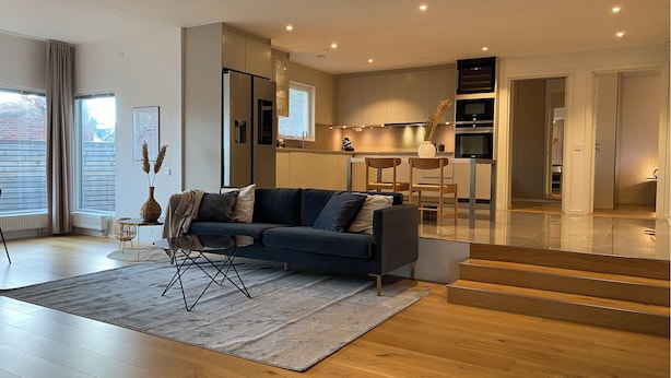 A modern living room with a sofa, a coffee table, and a rug. There is a kitchen in the background with cabinets and a dining area. - Exclusive newly apartment with free parking (Västra Götalands län)