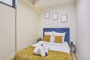 A neatly made bed with a blue headboard, white bedding, and a mustard-colored throw blanket. There are two bedside tables with small potted plants. - Apartment Exposition Center (Paris)