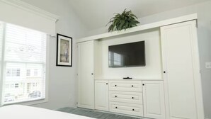 A bedroom with a bed, a TV mounted on a white cabinet, a window with blinds, and a plant on top of the cabinet. - Enjoy Every Moment in a Space Made for Great Getaways! 3 Units, Pool, Parking! (Williamsburg)