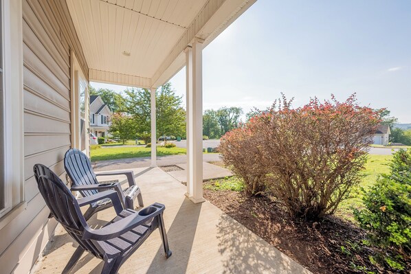 A covered porch with two Adirondack chairs, a small garden with a bush, and a view of a residential street.