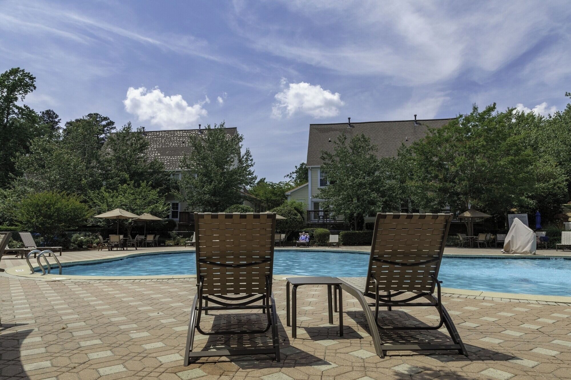A pool area with two lounge chairs and a small table, surrounded by a brick patio and trees.