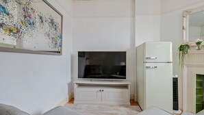 A living room with a flat-screen TV on a white entertainment unit, a white refrigerator, and a green radiator.