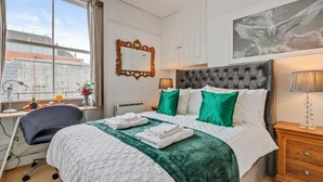 A hotel room with a large bed, a desk with a chair, a window with curtains, a radiator, a framed mirror, a nightstand with a lamp, and a painting on the wall.