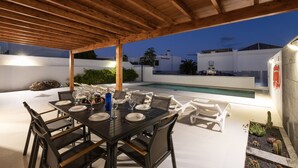 An outdoor dining area with a wooden pergola, a pool, and white walls.