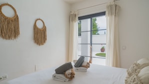 Een minimalistische slaapkamer met een groot bed, twee macramé wanddecoraties en uitzicht op groen buiten.