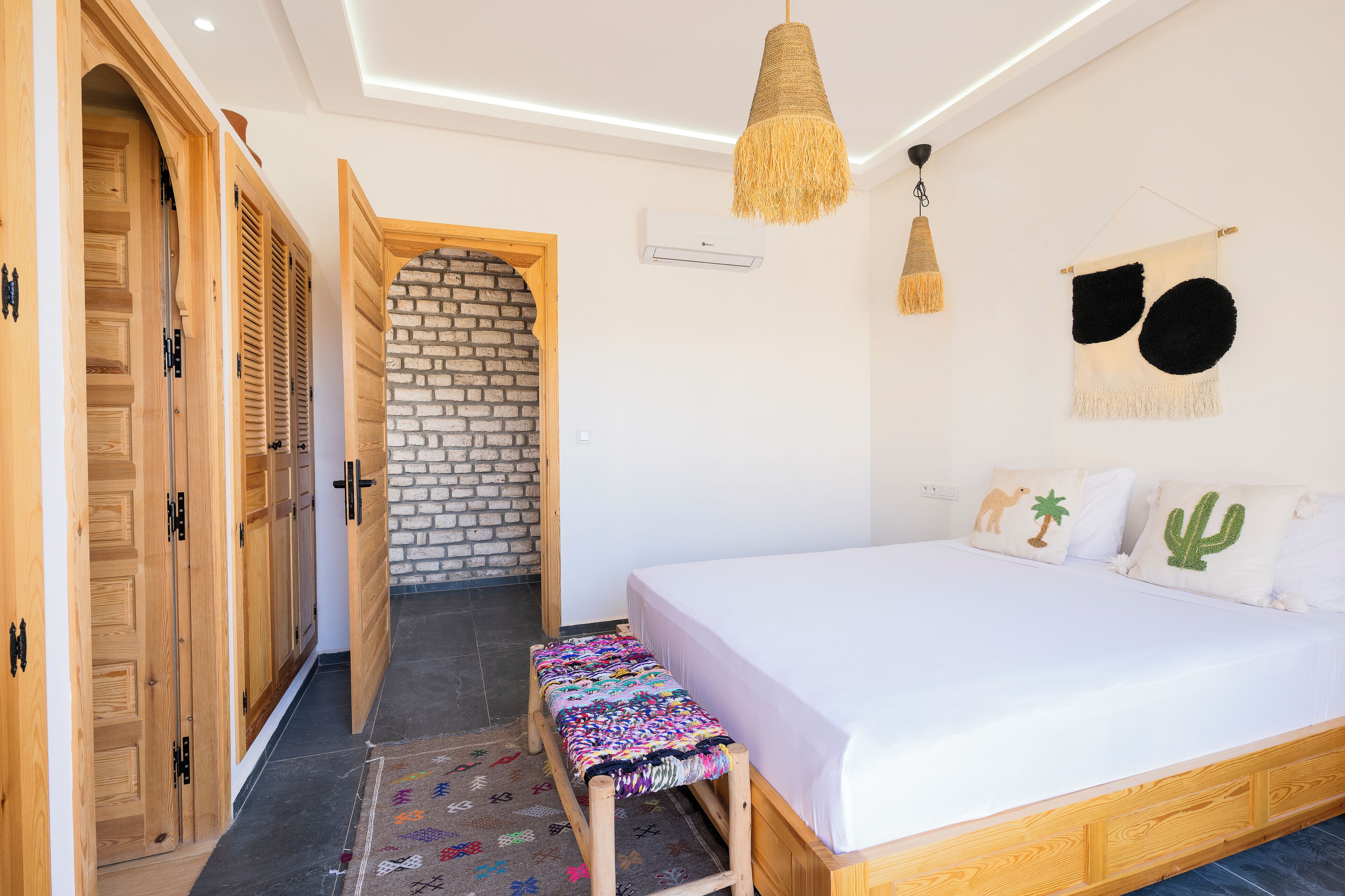 A bedroom with a bed, wooden wardrobe, bench, and a brick wall.