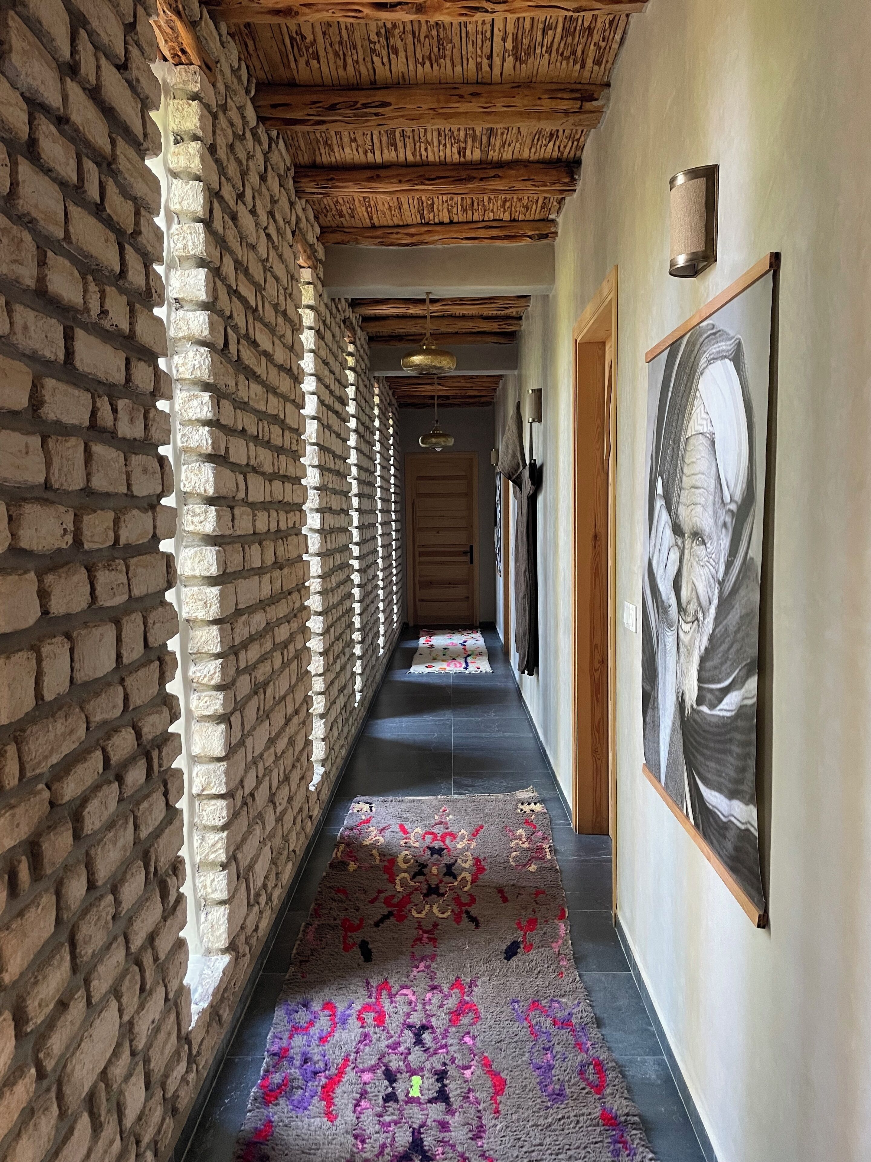 A corridor with a brick wall, wooden ceiling beams, a patterned carpet, and a framed artwork on the wall.