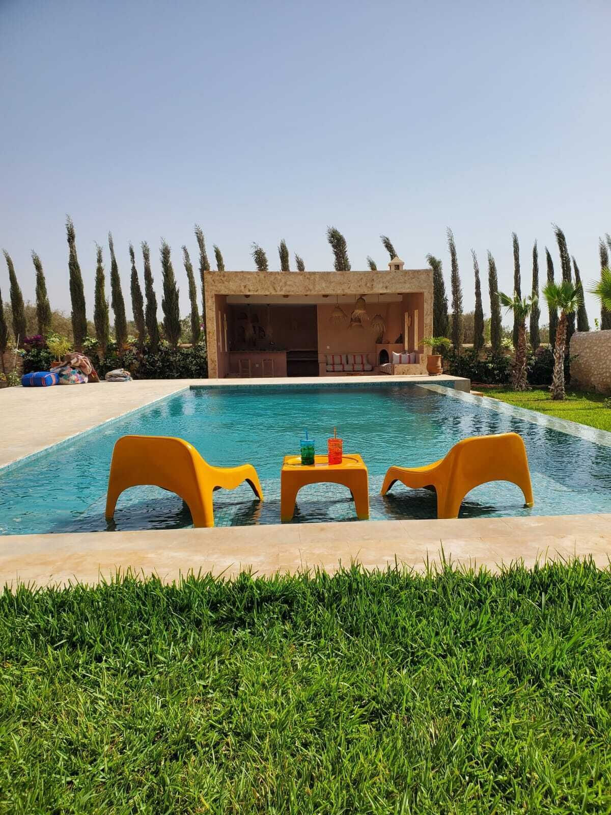 A swimming pool with two yellow chairs and a table, surrounded by a well-maintained lawn and tall trees.