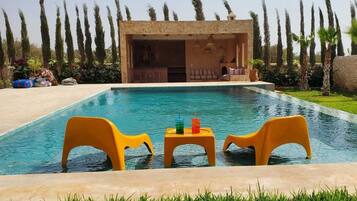 A swimming pool with two yellow chairs and a table, surrounded by a well-maintained lawn and tall trees.