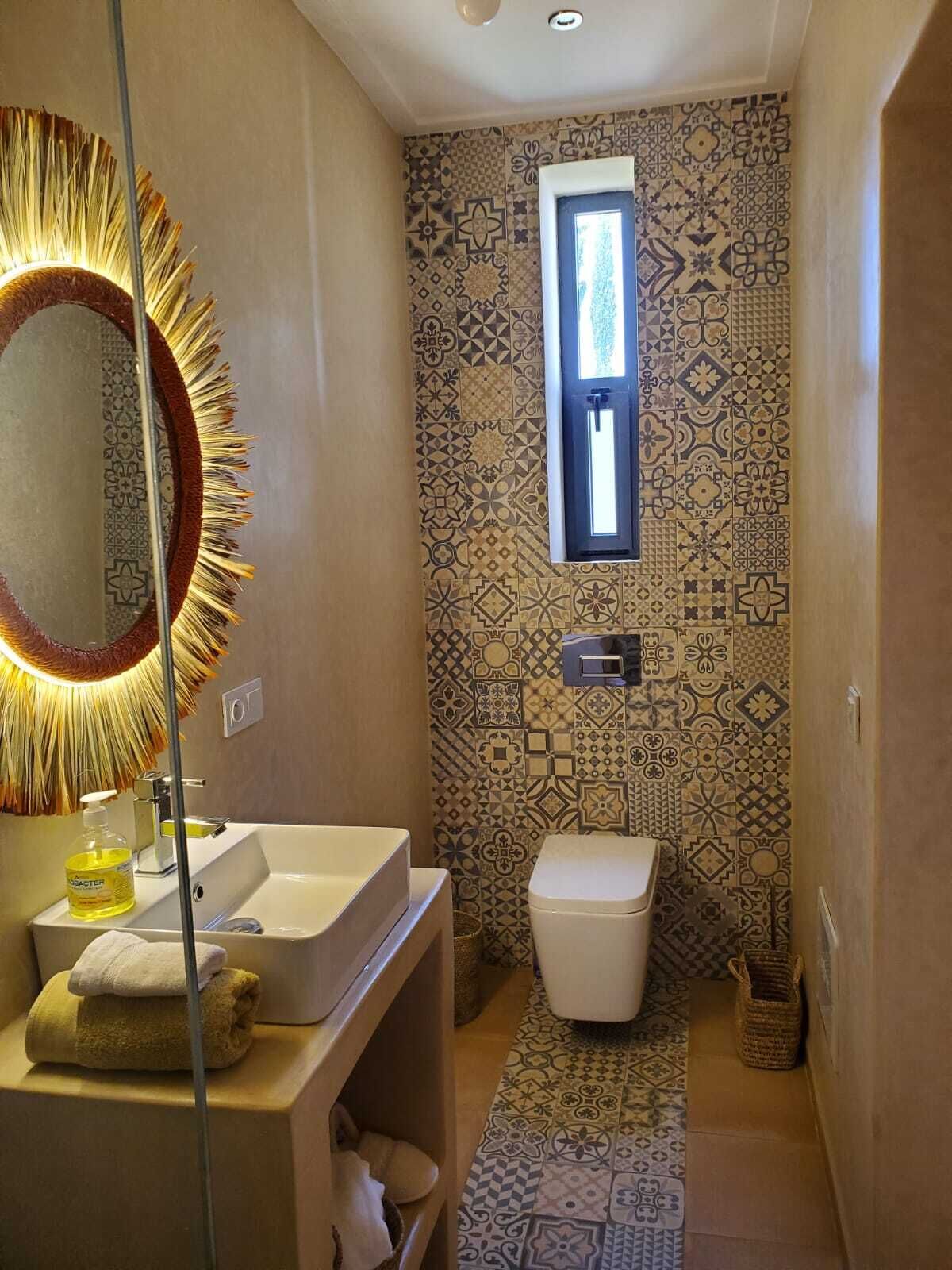 Et badeværelse med mønstret fliser, et spejl med solstråle-ramme, en vask, et toilet og et lille vindue.