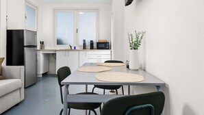 A compact kitchen with white cabinets, a refrigerator, a microwave, and a small countertop. There is a dining table with chairs and a potted plant on the wall.