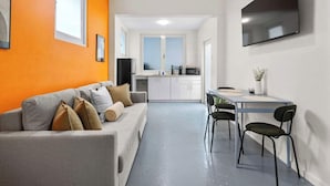 A modern living room with a grey sofa and orange walls, a dining area with a table and chairs, and a kitchen in the background.
