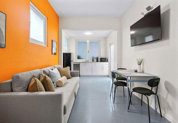 A modern living room with a grey sofa and orange walls, a dining area with a table and chairs, and a kitchen in the background.