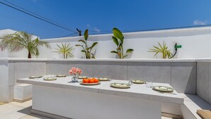 Un área de comedor al aire libre con una mesa blanca dispuesta para seis personas, rodeada de plantas en macetas.