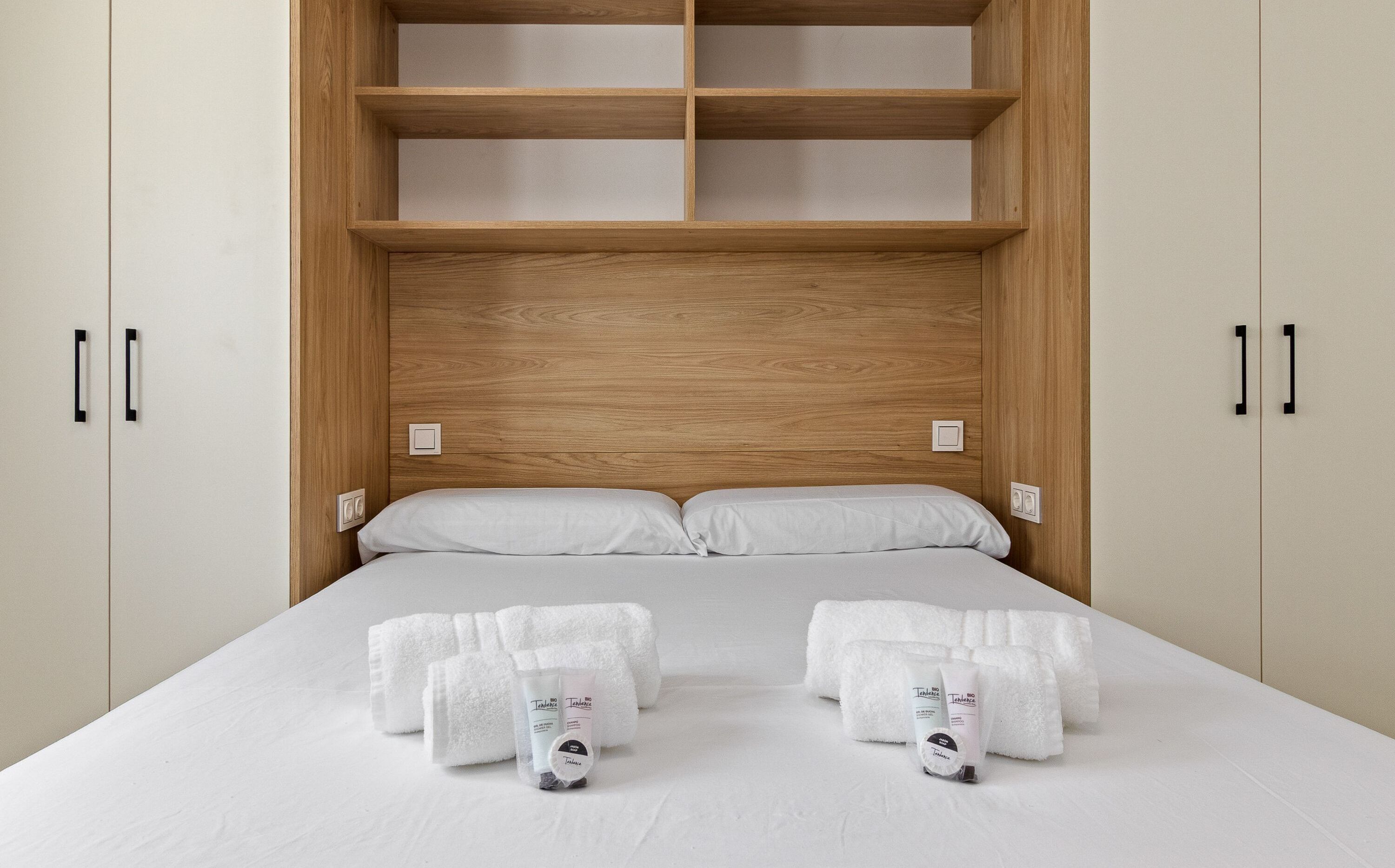 A bedroom with a wooden headboard, a bed with white linens, two towels, and toiletries.
