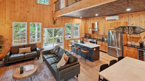 Amplia sala de estar con sofá, mesa de centro y mesa de comedor. Cocina con refrigerador, estufa y taburetes de bar. Ventanas amplias ofrecen vistas del bosque circundante.
