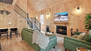 A cozy wooden interior with a green sofa, a dining area, a fireplace, and a TV displaying a snowy scene.