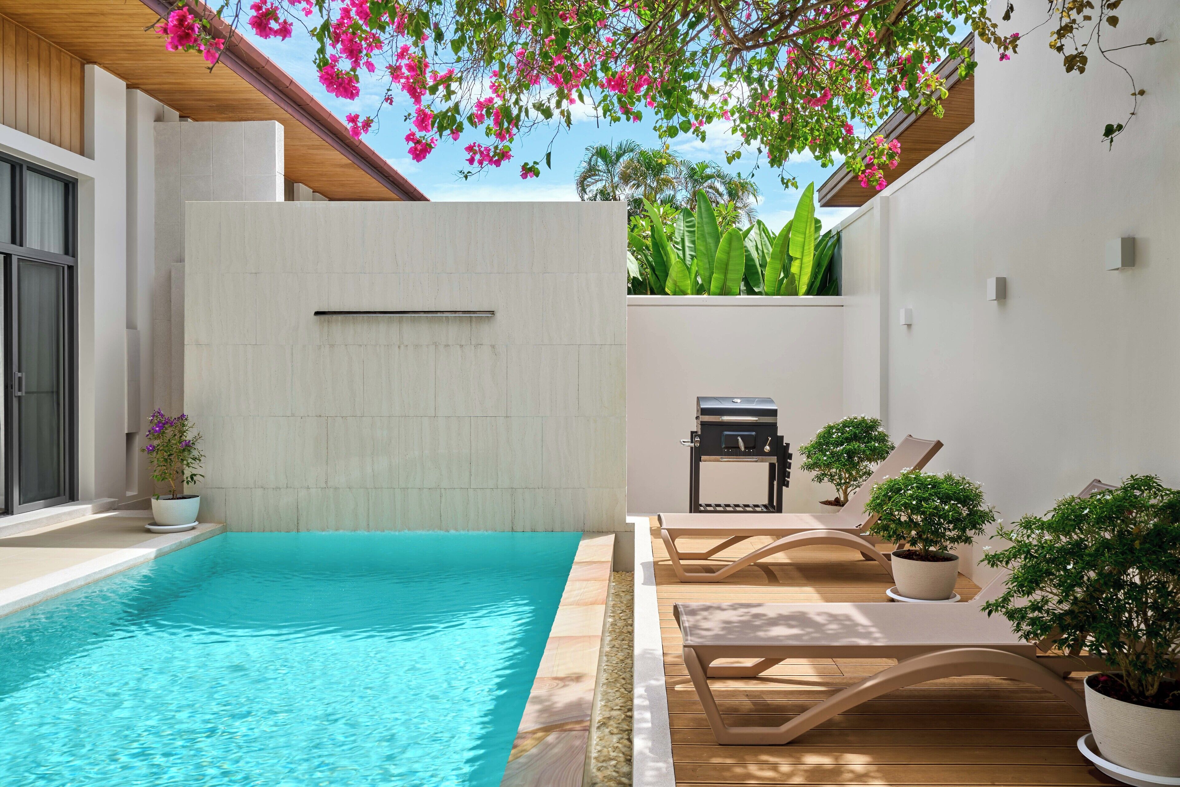 A swimming pool with lounge chairs and a barbecue grill, surrounded by potted plants and a wooden ceiling.