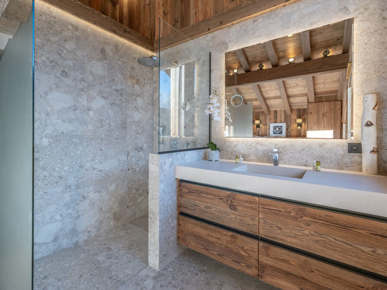 A bathroom with a stone wall, wooden cabinets, a glass shower enclosure, and a large mirror.