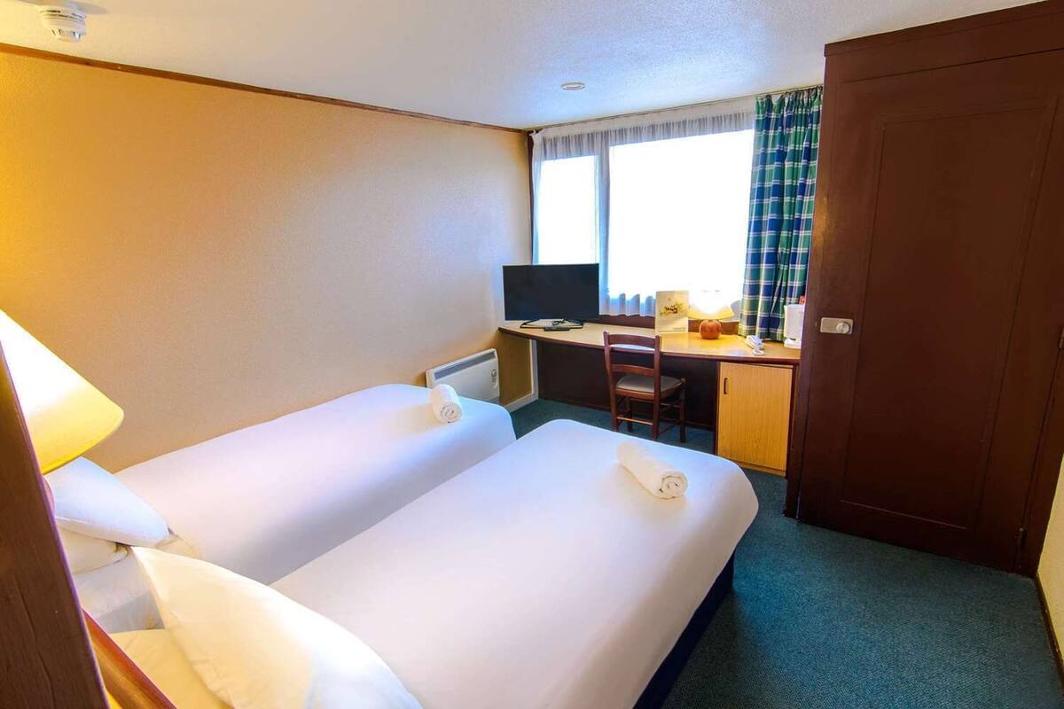 A hotel room with two beds, a desk with a computer, a TV, and a window with curtains.