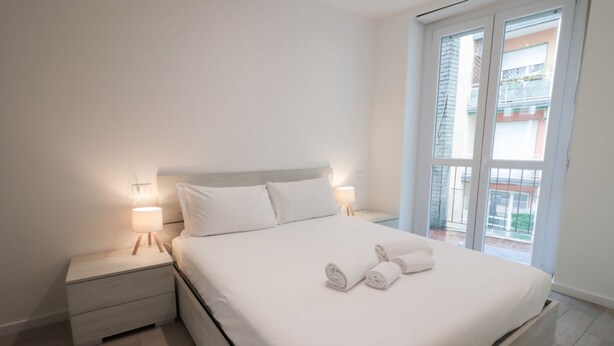 Una cama bien tendida con sábanas blancas y dos toallas enrolladas, una mesita de noche con una lámpara y una ventana con vistas a edificios.